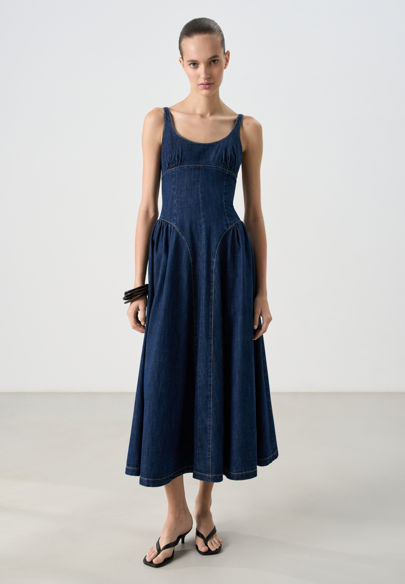 Джинсовое платье длины миди из 100% хлопка. Модель приталенного силуэта с акцентными вытачками по фигуре, без рукавов, U-образным вырезом, на тонких нерегулируемых бретельках и расклешенной юбкой. Застежка на молнию сзади.