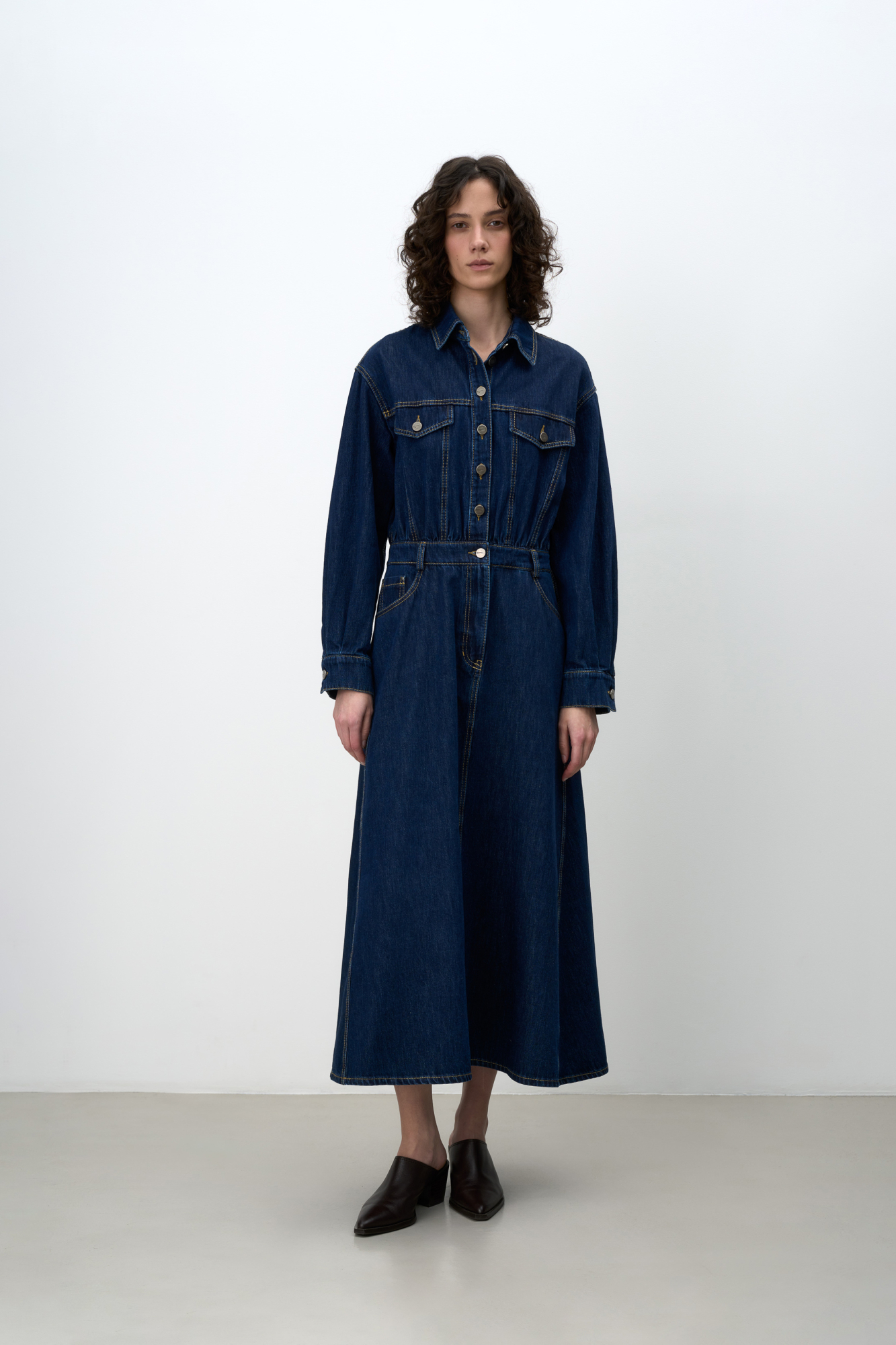 Джинсовое платье из 100% хлопка с рубашечнымм верхом и юбкой А-силуэта. Модель с длинными рукавами с манжетами, отложным воротником, карманами на груди с клапанами, шлевками на поясе для ремня, боковыми и задними карманами. Застежка на металлические пуговицы. Изделие декорировано вытачками контрастного цвета.