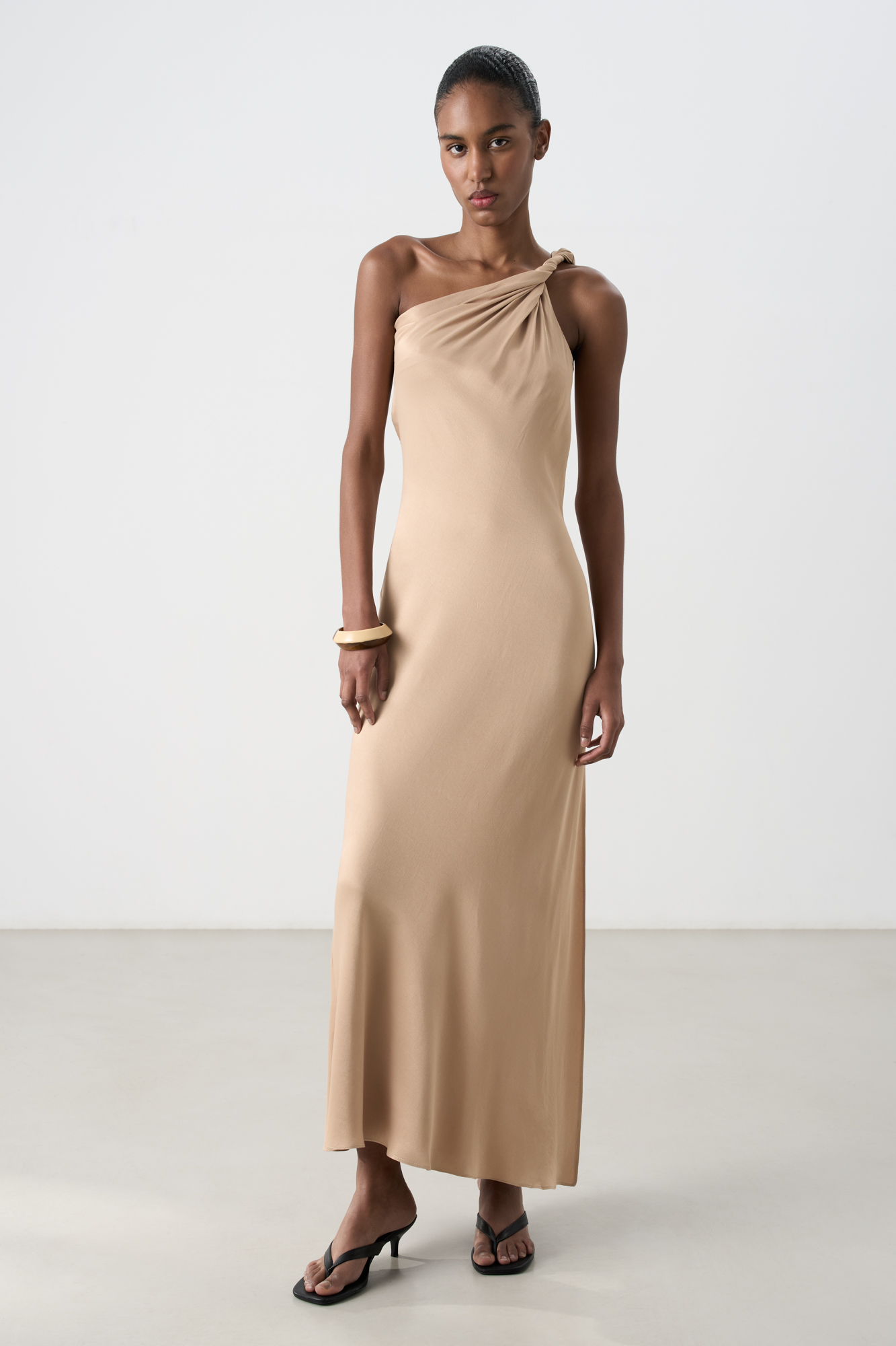 Платье длины макси из атласной вискозы. Модель приталенного силуэта, без рукавов, с перекрученной лямкой на одно плечо и разрезом сбоку.
