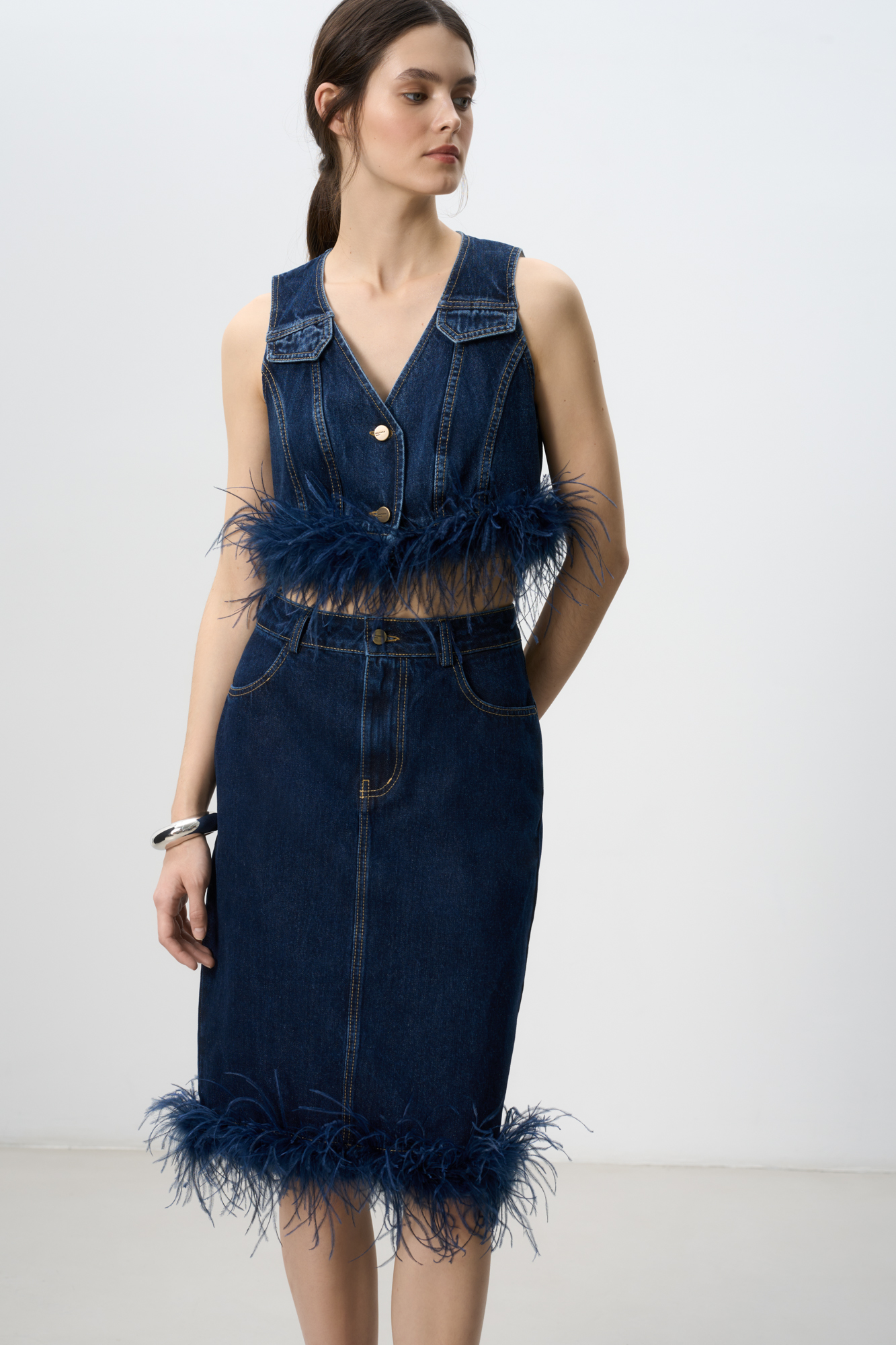 Джинсовая юбка длины миди, декорированная лентой со страусиными перьями. Лента с перьями отстегивается. Застежка изделия на молнию и пуговицу. Два кармана, шлевки на поясе для ремня.