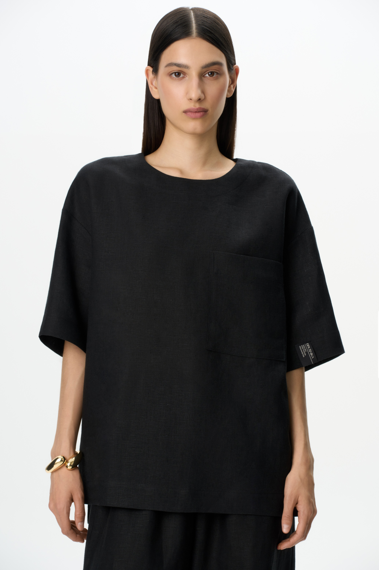 Широкая футболка из 100% льна. Модель прямого кроя, со спущенной линией плеча, круглым вырезом горловины и асимметричным низом с разрезами по бокам. Изделие декорировано накладным карманом на груди и брендированным ярлыком на рукаве. Застежка на пуговицу сзади.