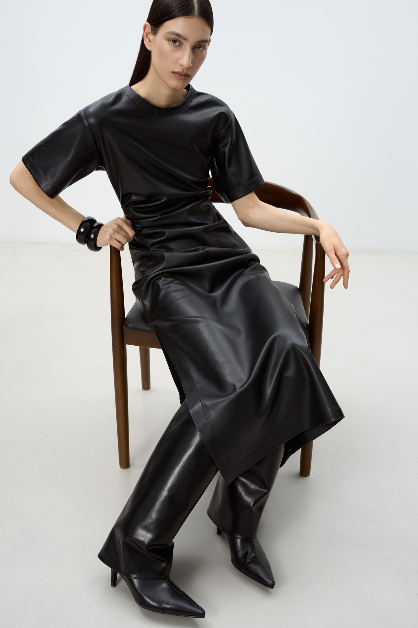 Длинное платье из экокожи. Модель свободного силуэта с коротким рукавом и круглым вырезом горловины. Изделие декорировано драпировкой и разрезом сбоку.