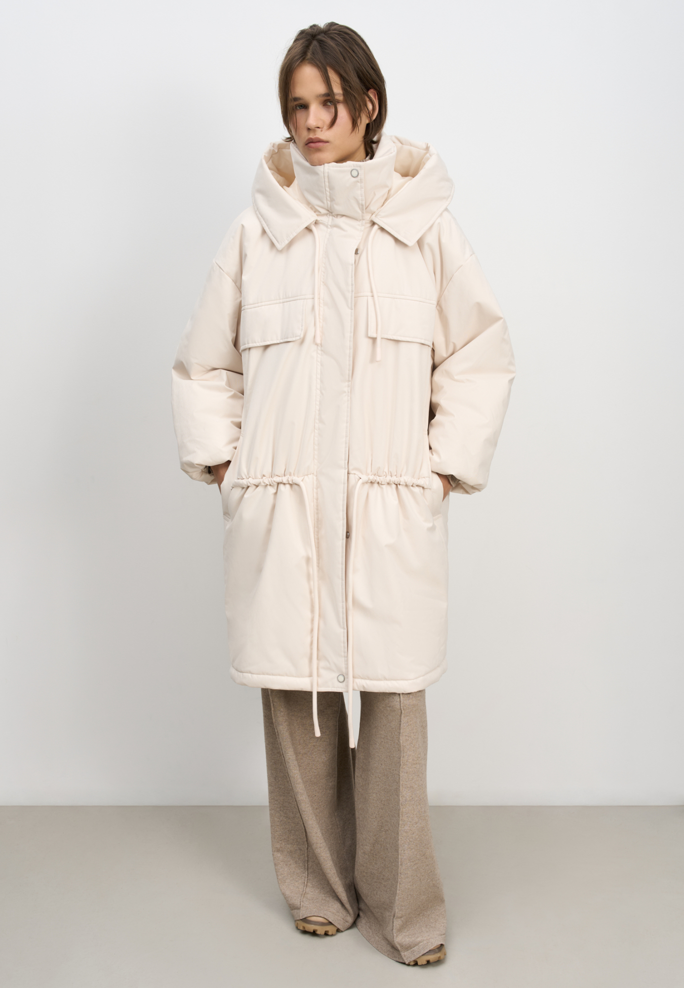 Длинная куртка на синтепоне с капюшоном. Модель свободного силуэта со спущенной линией плеча, накладными и боковыми карманами, регулируемой кулиской на поясе. Изделие на подкладке. Застежка на молнию и магнитные кнопки.