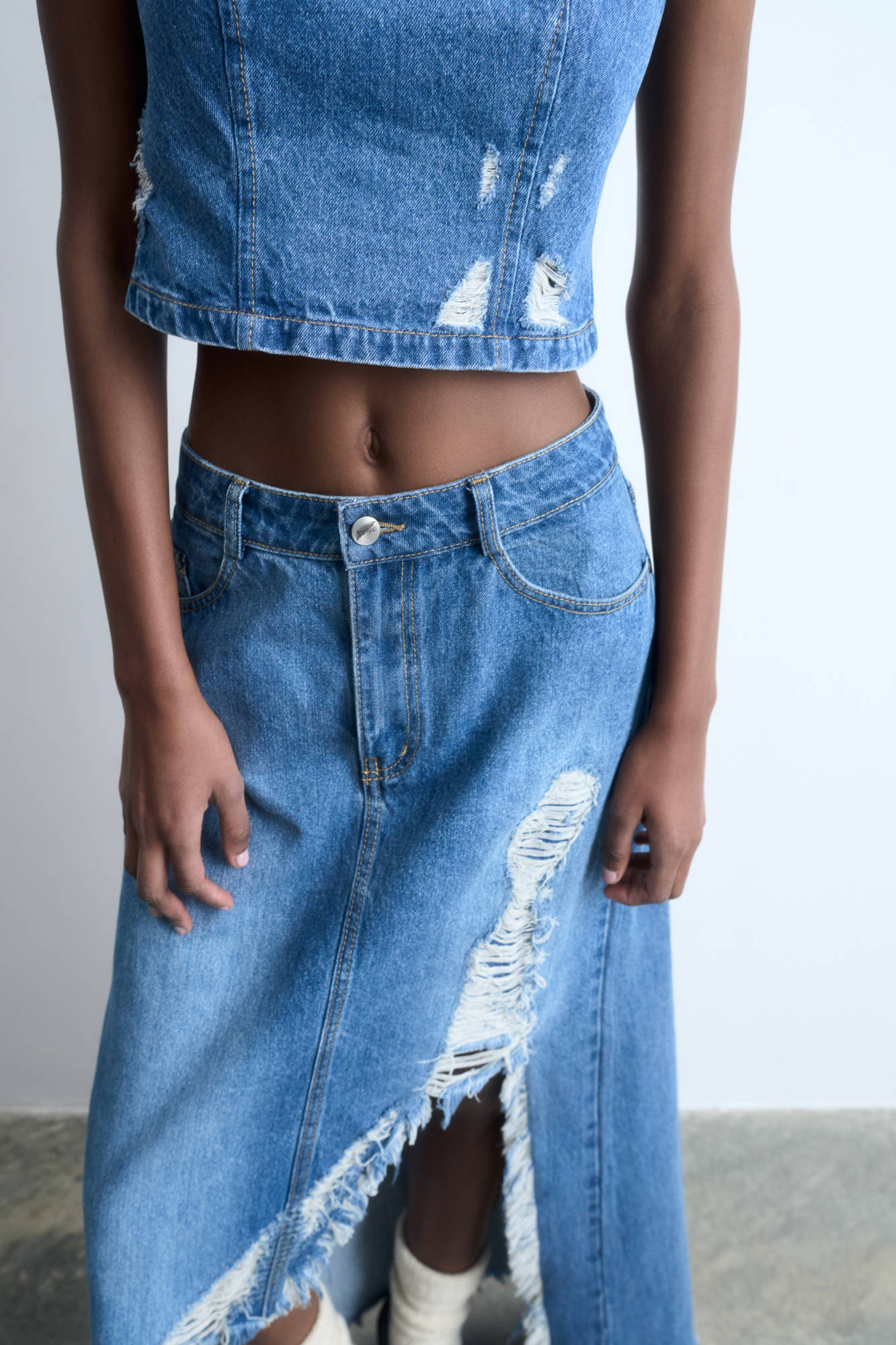 Джинсовая юбка из 100% хлопка в стиле гранж. Модель прямого кроя длины миди, со средней посадкой, пятью карманами и шлевками на поясе для ремня. Застежка на молнию и пуговицу. Изделие декорировано рваным разрезом от бедра, создающим асимметрию юбки.