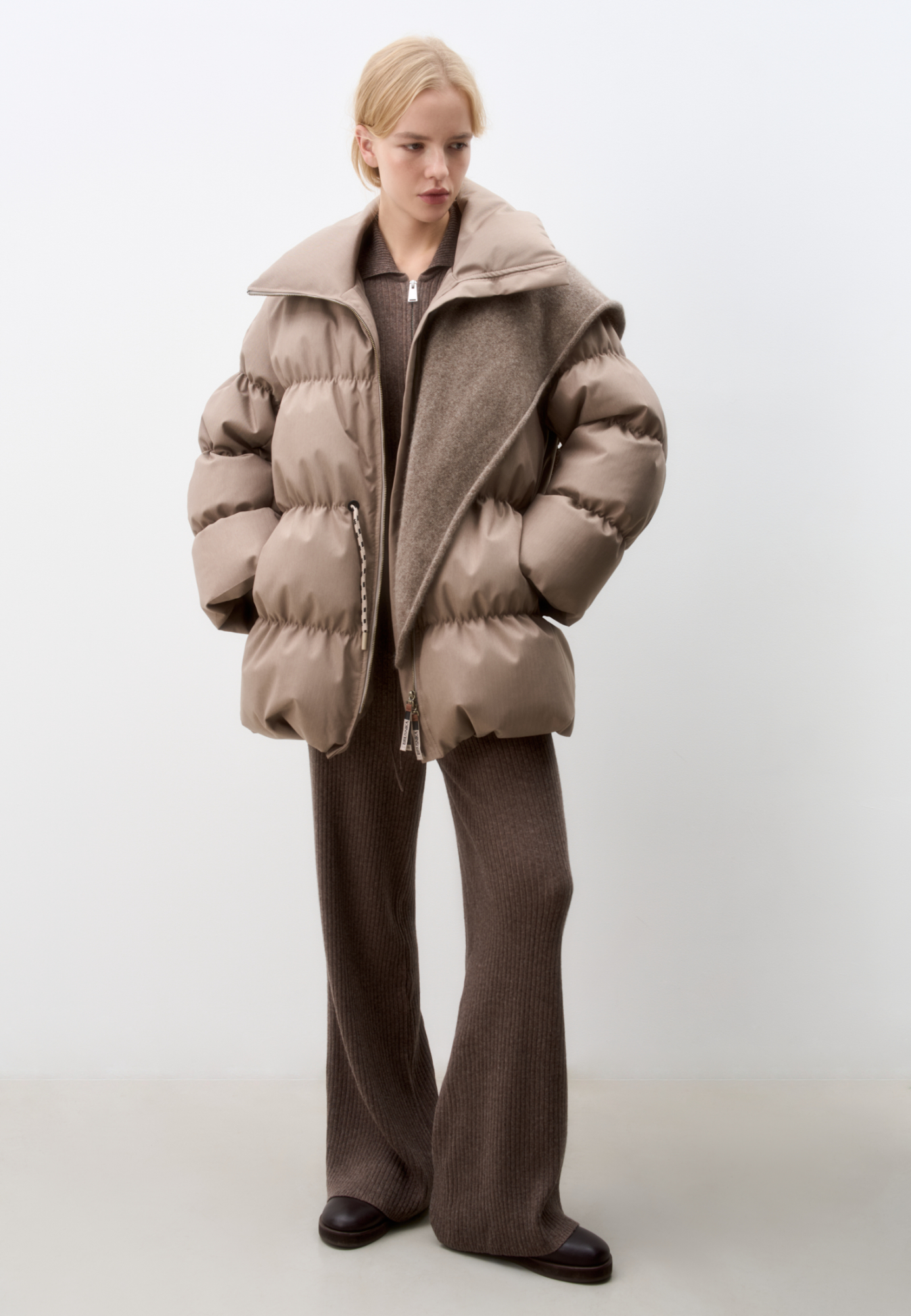 Стеганый объемный пуховик. Модель свободного силуэта, с воротником-стойкой, кулиской на талии и логотипом бренда на спине. Застежка на молнию. 