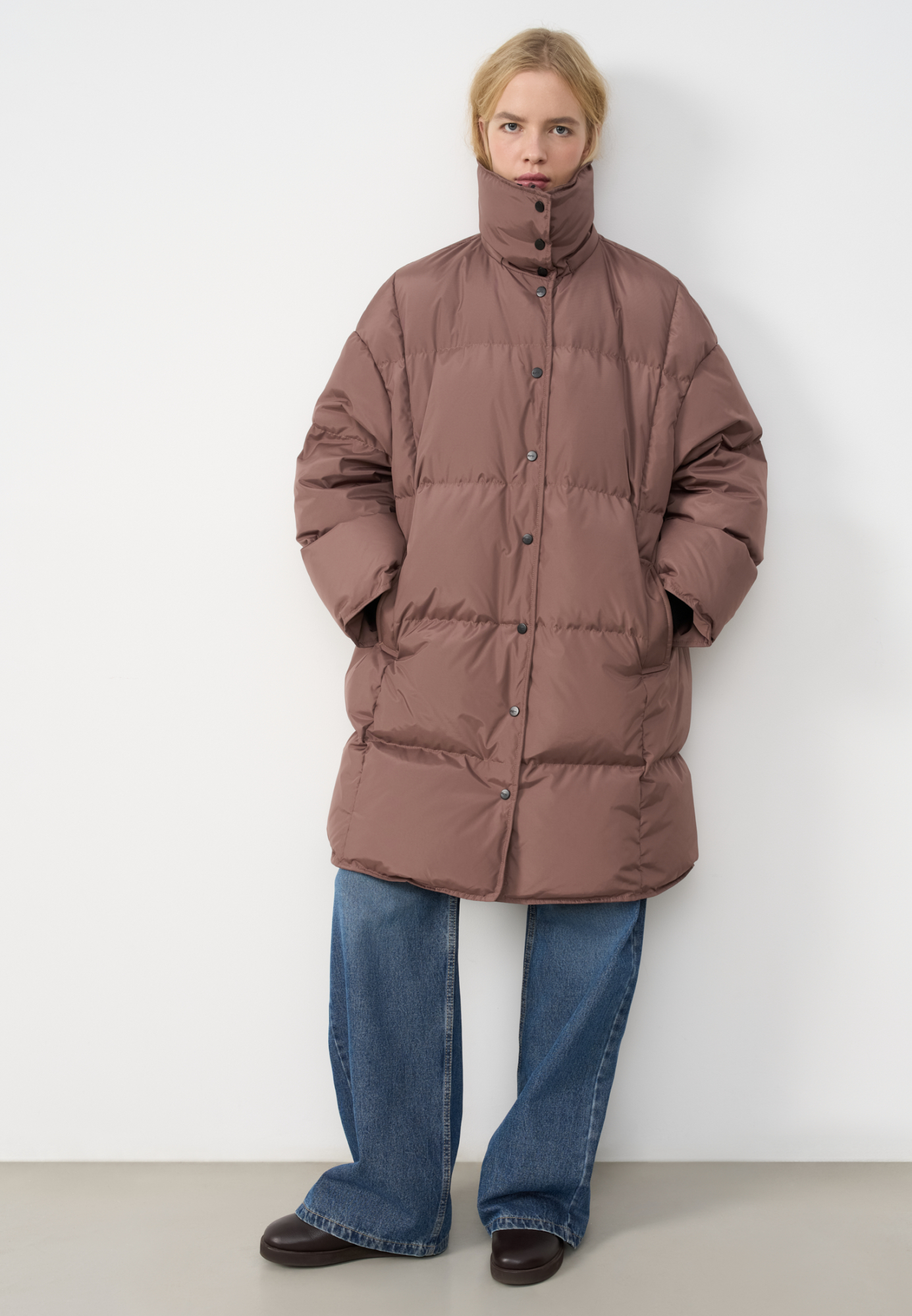 Длинный стеганый пуховик с капюшоном на кулиске. Модель свободного силуэта с воротником-стойкой, трикотажными манжетами и боковыми карманами. Застежка на молнию под планкой с брендированными пуговицами.