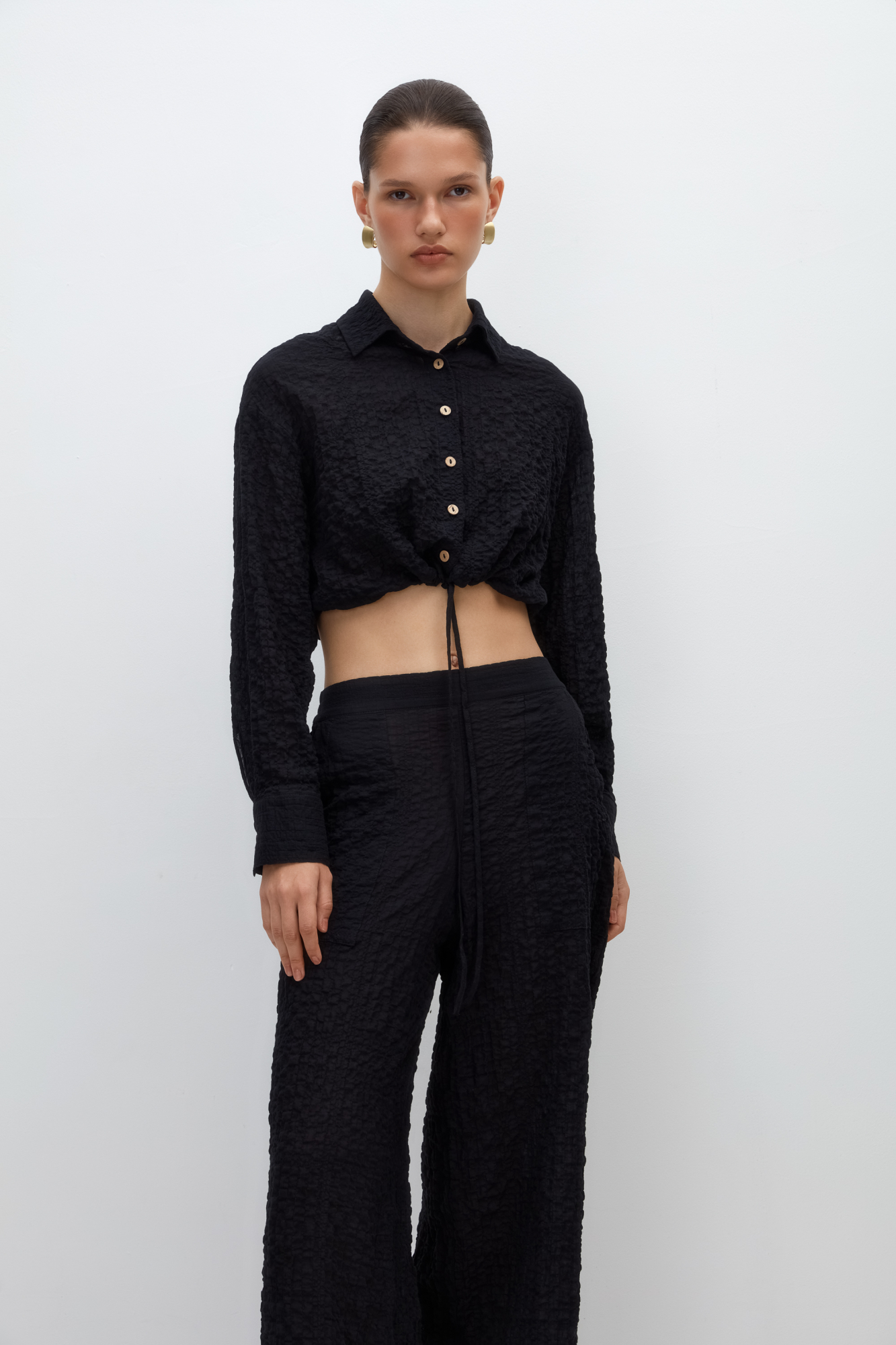 Укороченная рубашка из текстурной марлевки. Модель прямого кроя, с длинными рукавами, классическим отложным воротником, кулиской по низу и застежкой на пуговицы. 