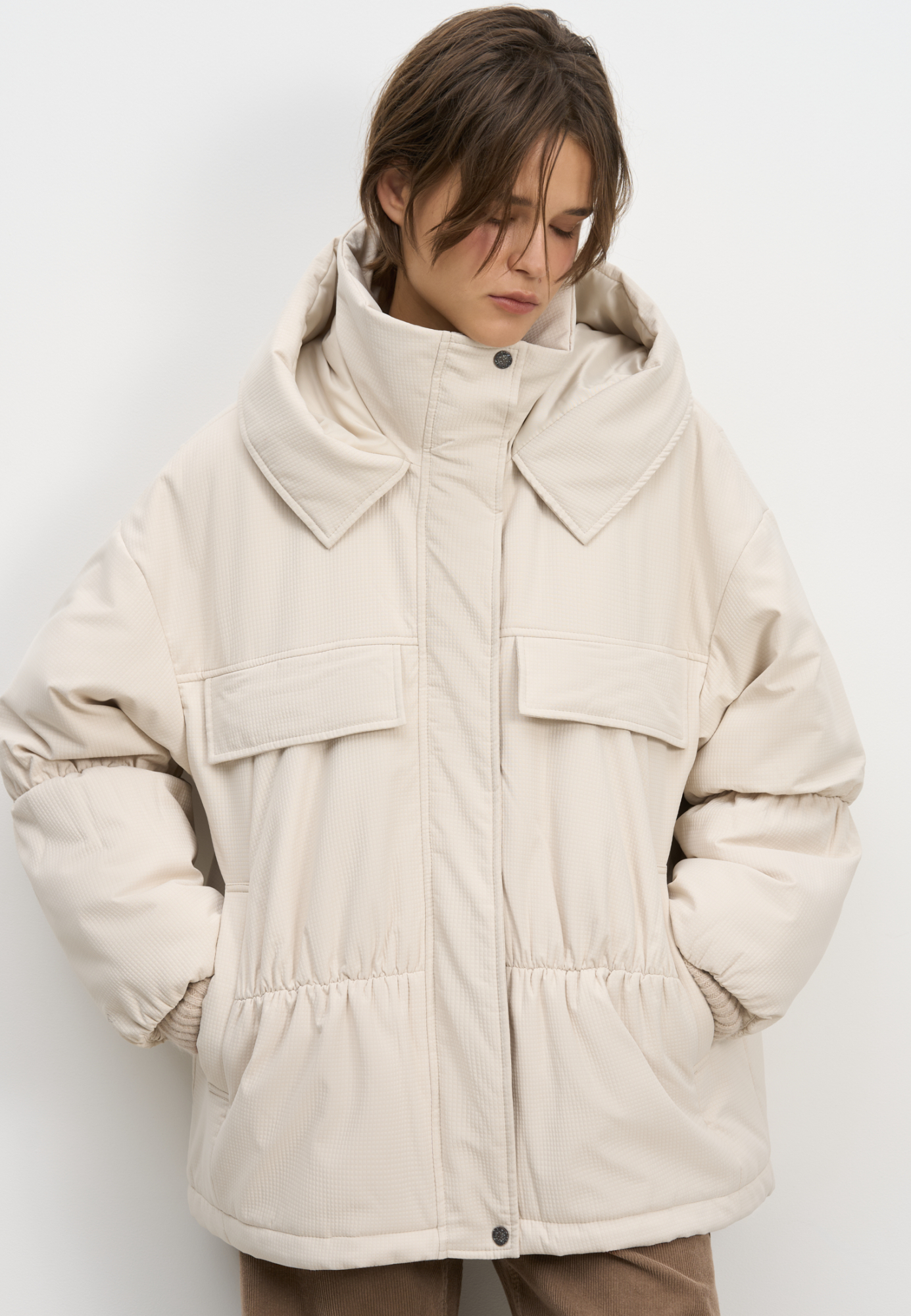 Длинная куртка на синтепоне из текстурной ткани. Модель свободного силуэта со спущенной линией плеча, накладными и боковыми карманами, воротником-стойкой и капюшоном. Изделие на подкладке. Застежка на молнию под планкой с кнопками.